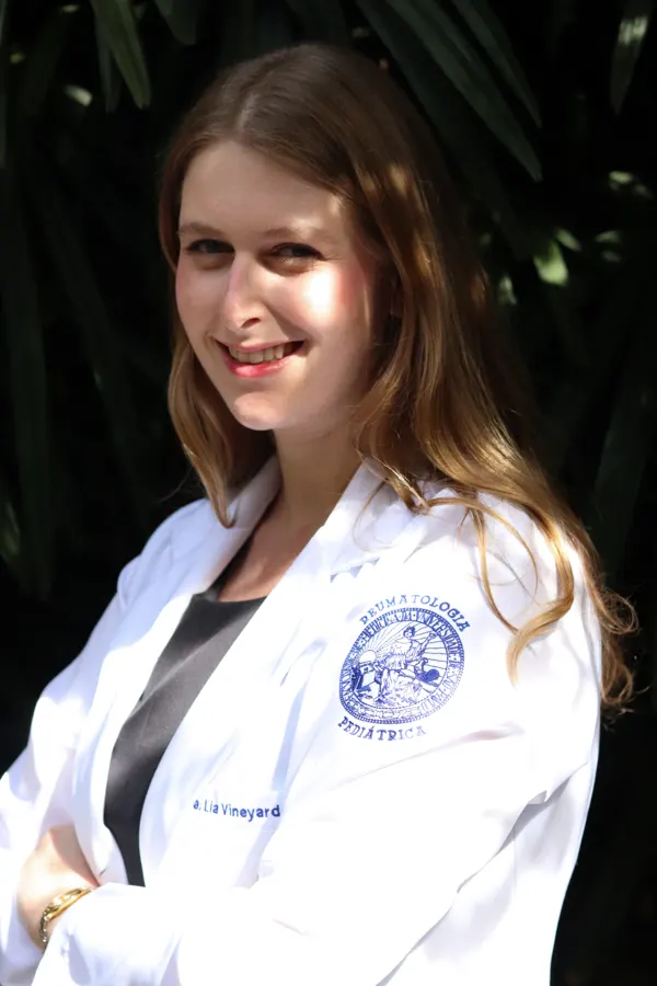 Dra. Lia Steuer de jaleco, sorrindo, em frente a folhagens